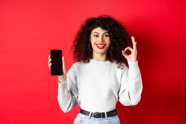 Fröhliche europäische Frau mit lockigem Haar, die einen leeren Handy-Bildschirm und eine gute Geste zeigt, zufrieden lächelt, gute App oder Werbung lobt, roter Hintergrund