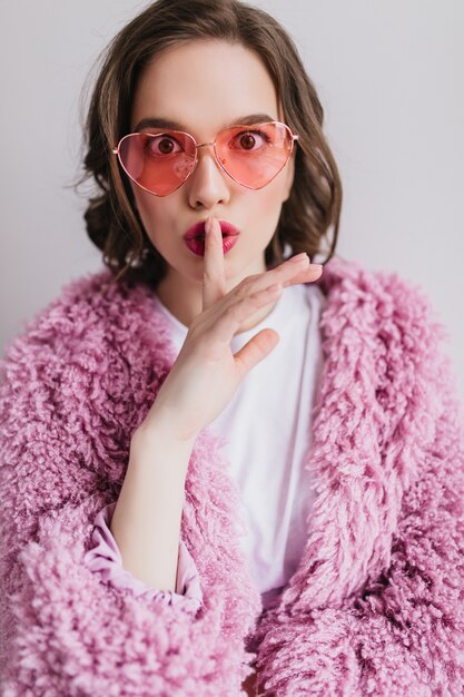 Fröhliche europäische junge Frau im trendigen flauschigen Mantel, der ihre Lippen mit Finger berührt. erstaunliches anmutiges Mädchen in der Sonnenbrille lokalisiert auf weißer Wand.