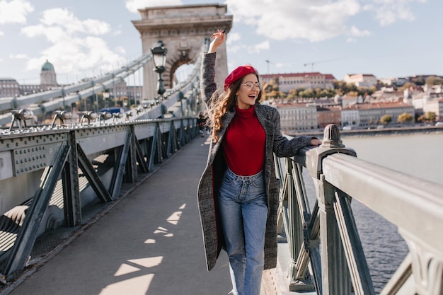 Fröhliche französische Reisende im roten Barett mit Blick auf den Fluss von der Brücke. Outdoor-Porträt einer entspannten jungen Dame in Vintage Blue Jeans, die einen Wochenendspaziergang durch die Stadt genießt.