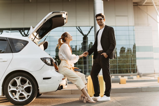 Fröhliche Frau in beigefarbener Hose mit weißer Bluse sitzt im Auto und hält die Hand ihres Freundes Brünetter Mann in schwarzem Anzug und Brille lächelt und posiert in der Nähe des Flughafens