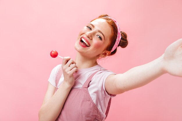 Fröhliche Frau in rosa Overall und weißem Oberteil hält Süßigkeiten und macht Selfie auf rosa Hintergrund.