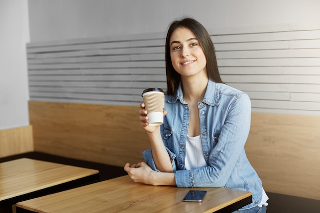 Fröhliche frau mit dunklem haar in trendigen kleidern, die in der cafeteria sitzt, nach einem langen arbeitstag kaffee trinkt, verträumt beiseite schaut und über dinge nachdenkt, die sie heute getan hat. lifestyle-konzept.