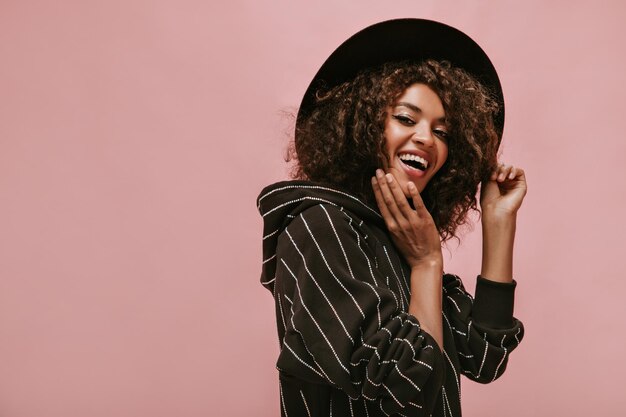 Fröhliche Frau mit dunkler, gewellter Frisur in schwarz gestreiftem Outfit und Hut, die lacht und auf rosa Hintergrund in die Kamera schaut