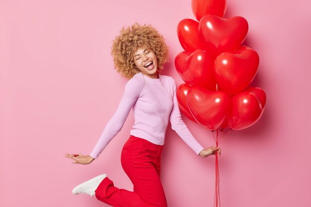 Fröhliche Frau mit lockigem Haar in lässigem Pullover und roter Hose hält einen Haufen Herzballons feiert besonderen Anlass fühlt sich sehr froh isoliert über rosa Hintergrund Urlaubskonzept