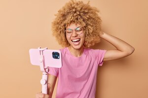 Kostenloses Foto fröhliche frau mit lockigen haaren verwendet selfie-stick und smartphone zum aufnehmen von bildern. sie lacht glücklich und trägt eine brille. lässiges rosafarbenes t-shirt, das über beigem hintergrund isoliert ist