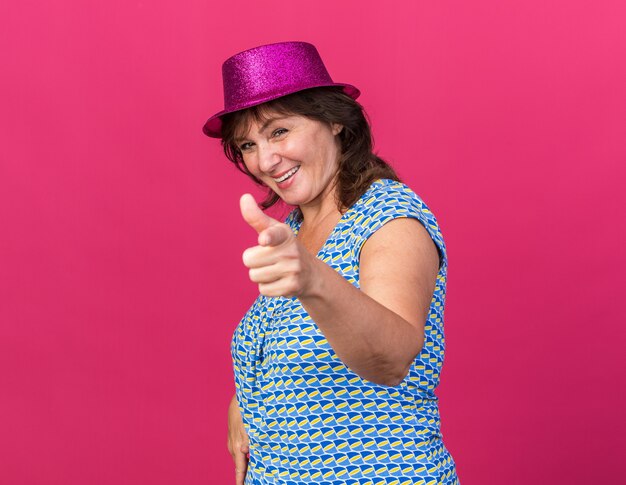 Fröhliche Frau mittleren Alters in Partyhut, die mit dem Zeigefinger zeigt, der fröhlich lächelt und die Geburtstagsfeier feiert, die über rosa Wand steht