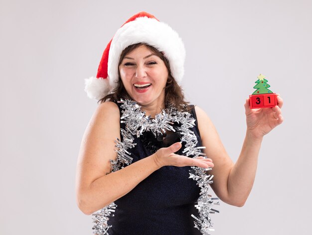 Fröhliche Frau mittleren Alters mit Weihnachtsmütze und Lametta-Girlande um den Hals, die auf Weihnachtsbaum-Spielzeug zeigt und mit Datum in die Kamera schaut, isoliert auf weißem Hintergrund