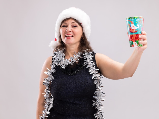 Fröhliche Frau mittleren Alters mit Weihnachtsmütze und Lametta-Girlande um den Hals, die einen Weihnachtsbecher aus Plastik in Richtung Kamera ausstreckt und auf die Kamera schaut, die auf weißem Hintergrund isoliert ist