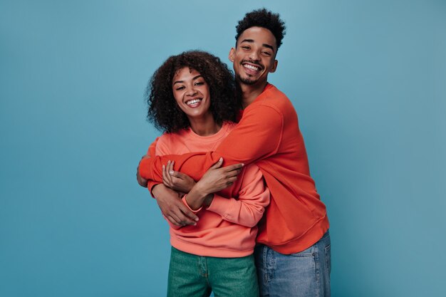 Fröhliche Frau und Mann in orangefarbenen Sweatshirts umarmen sich an der blauen Wand