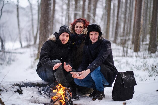 Fröhliche Freunde, die sich neben einem Lagerfeuer im kalten verschneiten Wald aufwärmen