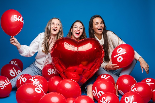 Fröhliche Freundinnen posieren mit rotem herzförmigem Ballon und Luftbällen mit Prozent- und Verkaufsbeschriftung auf blauem Hintergrund