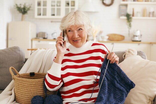 Fröhliche, freundlich aussehende, grauhaarige, ältere Frau in Freizeitkleidung, die auf einem Sofa mit Nadeln und einem Garten sitzt, ein nettes Telefongespräch mit ihrer alten Freundin führt, klatscht und die neuesten Nachrichten teilt