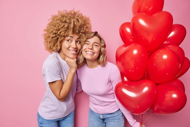 Fröhliche, freundliche Frauen haben frohe, verträumte Ausdrücke, die sich auf den Valentinstag vorbereiten. Stellen Sie sich eine tolle Party-Pose mit roten, herzförmigen Luftballons vor, die in lässige Kleidung gekleidet sind, die über rosa Hintergrund isoliert sind