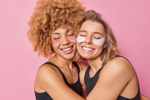 Fröhliche, freundliche Frauen umarmen sich mit Liebeslächeln und tragen sanft Schönheitsflecken unter den Augen auf, um die Haut zu behandeln und zu verjüngen, gekleidet in schwarzen T-Shirts, die über rosafarbenem Hintergrund isoliert sind