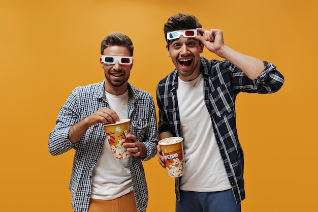 Fröhliche, fröhliche Brunet-Männer in weißen T-Shirts und blau karierten Hemden lächeln in 3D-Brillen, essen Popcorn und sehen sich einen Film auf orangefarbenem Hintergrund an