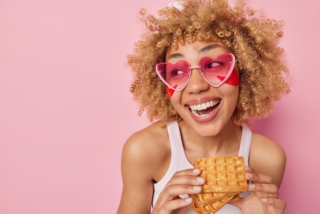 Fröhliche, fröhliche junge Frau hält köstliche, appetitanregende, hausgemachte Waffeln, lächelt weit weg, hat Naschkatzen, trägt Schönheitsflecken unter den Augen auf und trägt ein lässiges weißes T-Shirt mit rosa Sonnenbrille