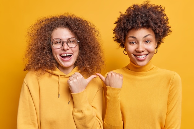 Kostenloses Foto fröhliche, fröhliche, lockige frauen mit gemischten rassen zeigen mit fröhlichem ausdruck aufeinander und sagen, dass sie eng beieinander stehen, lässig isoliert über gelber wand gekleidet. ich habe dich ausgewählt