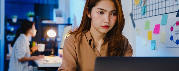 Fröhliche Geschäftsfrau aus Asien, die sich in einer neuen normalen Situation zur Virenprävention sozial distanziert, während sie in der Büronacht bei der Arbeit mit Laptop-Online-Geschäft Überstunden macht.