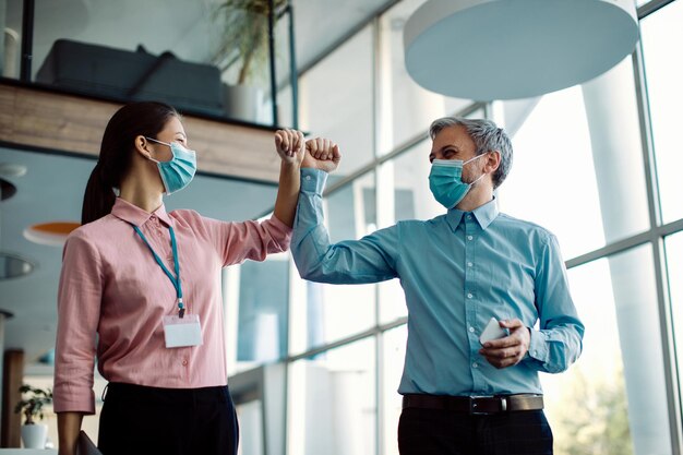 Fröhliche Geschäftskollegen schlagen sich mit dem Ellbogen und tragen Gesichtsmasken in einem Flur