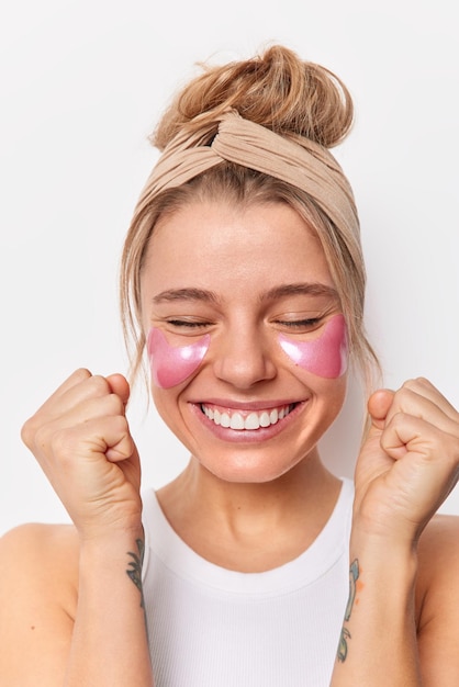 Fröhliche glückliche Frau ballt Fäuste freut sich über gute Nachrichten unterzieht sich Schönheitsbehandlungen wendet Hydrogel-Patches an genießt effektive Schönheitsbehandlung trägt beiges Stirnband isoliert über weißer Studiowand