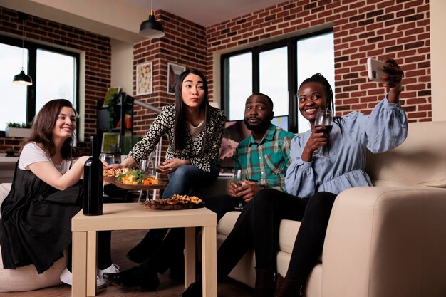 Fröhliche Gruppe von Freunden, die alkoholische Getränke trinken, während sie sich auf der Geburtstagsfeier im heimischen Wohnzimmer fotografieren. Verschiedene Menschen feiern ein Ereignis mit Essen und Wein, während sie Selfies machen.