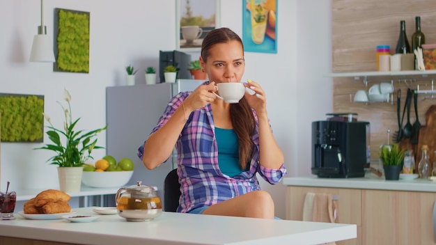 Fröhliche Hausfrau, die beim Frühstück aromatischen Tee trinkt. Frau, die einen großartigen Morgen hat, der leckeren natürlichen Kräutertee trinkt, der in der Küche sitzt und lächelt und Teetasse hält und mit angenehmen Erinnerungen genießt.