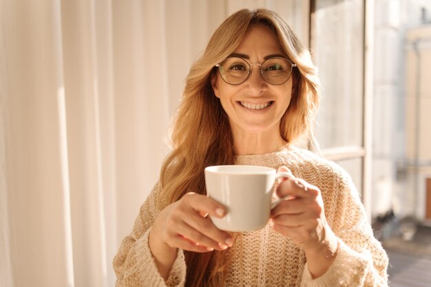Fröhliche hellhäutige erwachsene blonde Frau mit Brille, die in die Kamera lächelt, während sie aromatischen Kaffee trinkt, während sie am Fenster steht Freizeit, Entspannung und Lifestyle-Konzept