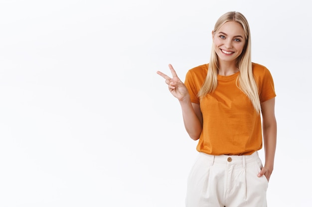 Fröhliche, hübsche, feminine, stilvolle blonde frau in orangefarbenen t-shirt-trendhosen, hand in der tasche halten, friedens- oder siegeszeichen als bescheidenes lächeln mit schönem, zufriedenem ausdruck zeigen, weißen hintergrund stehen