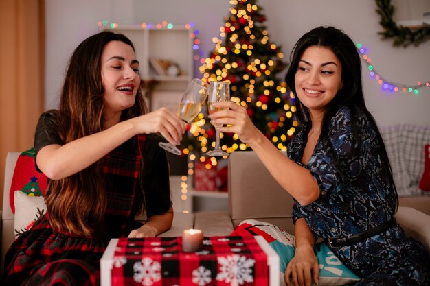Fröhliche hübsche junge Mädchen klirren und schauen auf Gläser Champagner, die auf Sesseln sitzen und die Weihnachtszeit zu Hause genießen