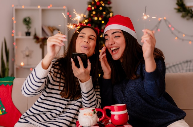 Fröhliche hübsche junge Mädchen mit Rentierbrille und Weihnachtsmütze halten Wunderkerzen und schauen auf das Telefon, das auf Sesseln sitzt und die Weihnachtszeit zu Hause genießt