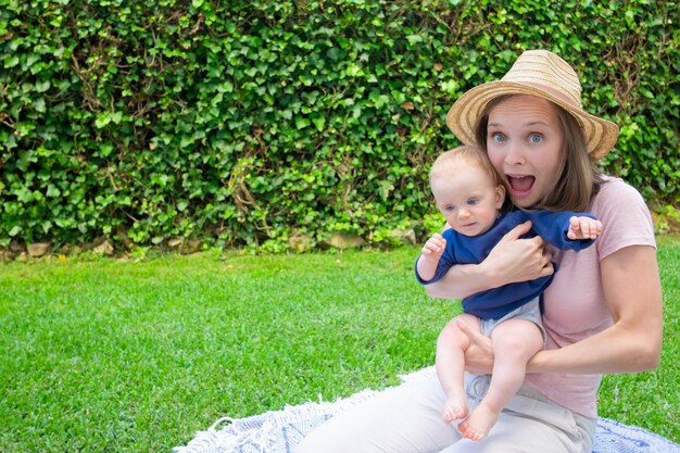 Fröhliche hübsche Mutter, die auf Plaid im Park mit offenem Mund sitzt, Neugeborenes hält und Kamera betrachtet. Nettes Baby im blauen Hemd auf Mutterhänden