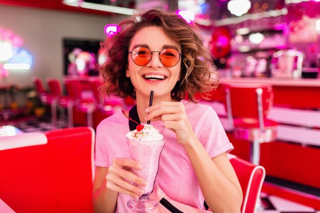 Fröhliche, hübsche, stilvolle lächelnde Frau im Retro-Vintage-Café der 50er Jahre, die am Tisch sitzt und Milchshake-Cocktail mit rosa Sonnenbrille trinkt und Spaß hat, mit offenem Lächeln zu lachen