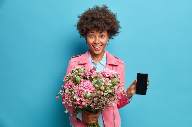 Fröhliche junge Afroamerikanerin hält Blumenstrauß zeigt Smartphone mit Modell Display lächelt positiv genießt besonderen Urlaub