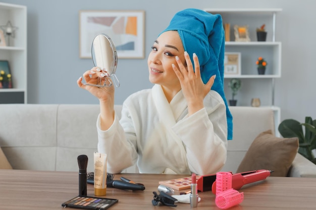 Fröhliche junge Asiatin mit Handtuch auf dem Kopf, die zu Hause am Schminktisch sitzt und einen Spiegel hält, der die morgendliche Make-up-Routine macht