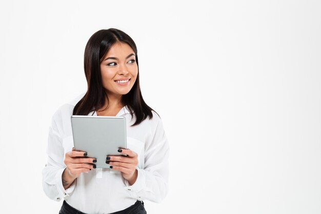 Fröhliche junge asiatische Dame, die durch Tablet-Computer plaudert. Zur Seite schauen.