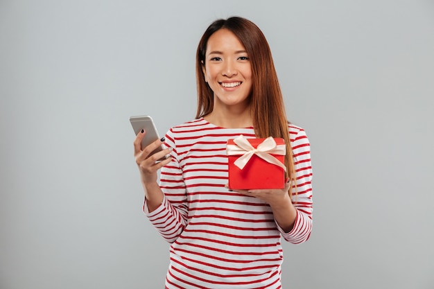 Fröhliche junge asiatische Frau, die per Telefon hält, das Geschenk hält.