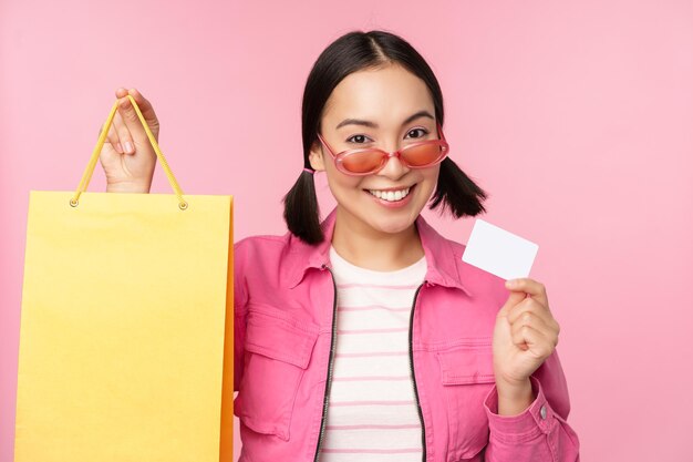 Fröhliche junge asiatische Frau zeigt Kreditkarten- und Einkaufstaschenverkaufsankündigung, die etw im Geschäft kauft, das vor rosafarbenem Hintergrund posiert