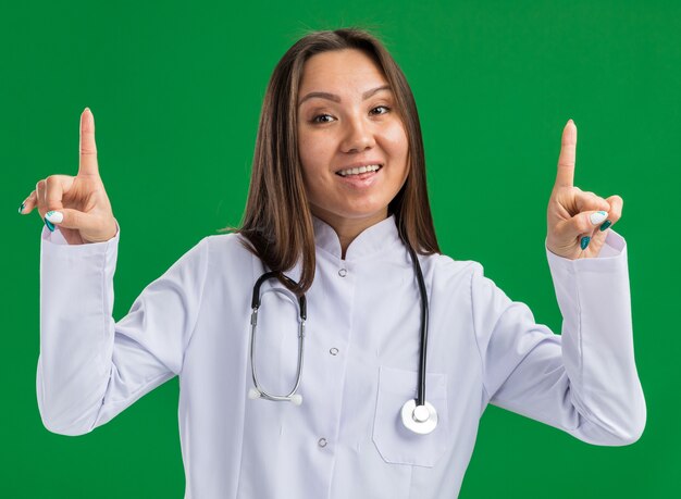 Fröhliche junge asiatische Ärztin mit medizinischem Gewand und Stethoskop, die nach vorne zeigt, isoliert auf grüner Wand