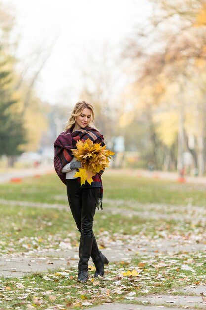 Fröhliche junge blonde Frau, die lächelt und einen Strauß gelber Herbstblätter anbietet. Schöne Dame, die in voller Länge im Park aufwirft.
