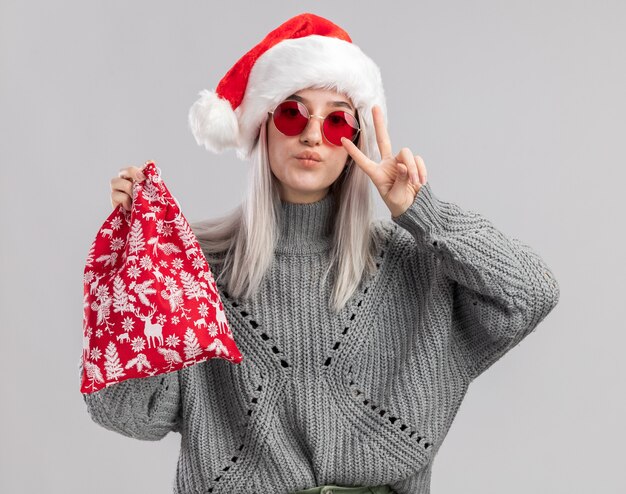 Fröhliche junge blonde Frau in Winterpullover und Weihnachtsmütze, die eine rote Weihnachtstasche mit Weihnachtsgeschenken mit einem V-Zeichen über der weißen Wand hält