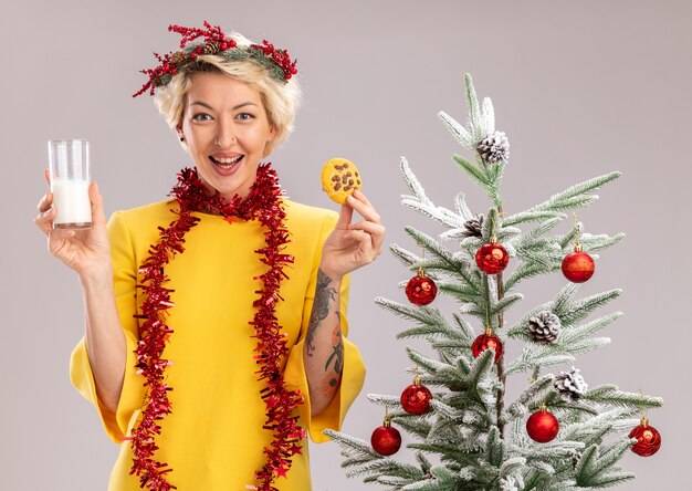 Fröhliche junge blonde Frau mit Weihnachtskopfkranz und Lamettagirlande um den Hals, die in der Nähe des geschmückten Weihnachtsbaums steht und ein Glas Milch und Kekse hält, die isoliert auf weißer Wand aussehen