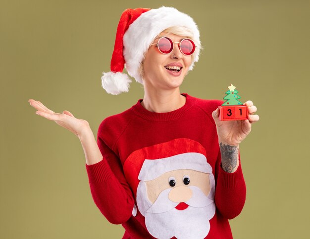 Fröhliche junge blonde Frau mit Weihnachtsmütze und Weihnachtsmann-Weihnachtspullover mit Brille mit Weihnachtsbaumspielzeug mit Datum, das leere Hand isoliert auf olivgrüner Wand zeigt