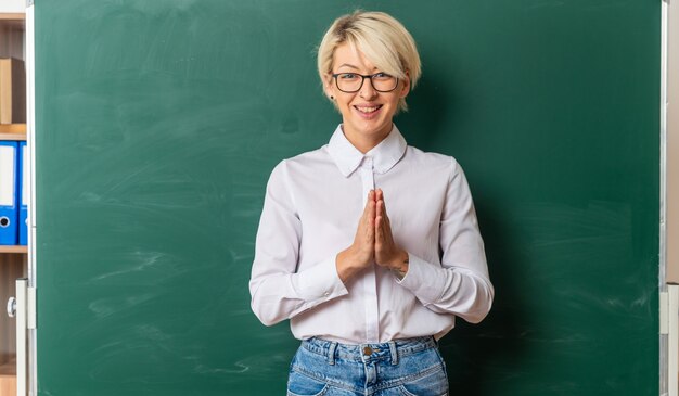 Fröhliche junge blonde Lehrerin mit Brille im Klassenzimmer, die vor der Tafel steht und die Hände zusammenhält und in die Kamera schaut