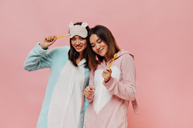 Fröhliche junge brünette asiatische Frauen in farbenfrohen, weichen Pyjamas lächeln aufrichtig und halten gelbe Zahnbürsten auf rosafarbenem Hintergrund