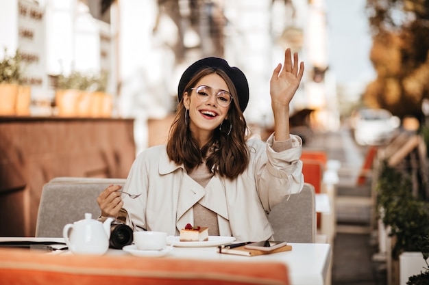 Fröhliche junge Brünette mit Baskenmütze, beigefarbenem Trenchcoat und stilvoller Brille, die am sonnigen Herbsttag auf der Terrasse des Stadtcafés sitzt, Käsekuchen isst und nach Kellner ruft