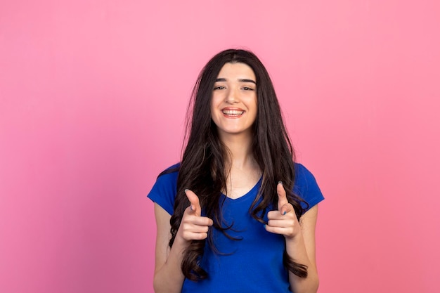 Fröhliche junge Dame, die ihre Finger auf die Kamera zeigt und auf rosafarbenem Hintergrund steht