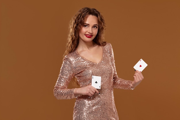 Kostenloses Foto fröhliche junge dame in einem goldglitzernden kleid hält zwei spielkarten in den händen, lächelt und blickt in die kamera. casino-konzept auf braunem hintergrund.