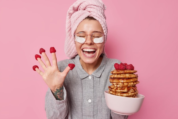 Fröhliche junge Europäerin isst köstliche Pfannkuchen mit Himbeerkichern und trägt positiv ein Nachtwäsche-Handtuch, das auf dem Kopf gewickelt ist, und eine transparente Brille, die über rosafarbenem Hintergrund isoliert ist Frühstückszeit