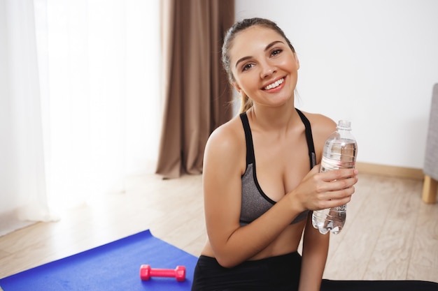 Fröhliche junge Fitness-Mädchen machen Yoga-Übungen auf dem Boden und trinken Wasser