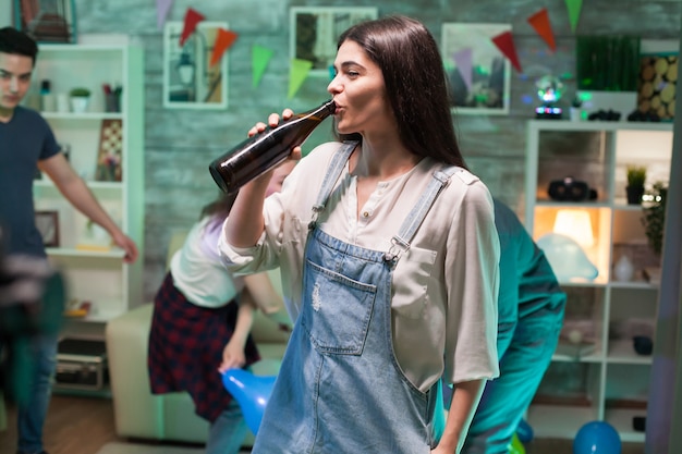 Fröhliche junge Frau auf einer Party mit ihren Freunden, die einen Schluck Bier trinken. Junge Leute tanzen.